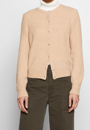 Femme portant un cardigan beige boutonné sur un col roulé blanc, associé à un pantalon taille haute vert olive avec des poches plaquées à l'avant.