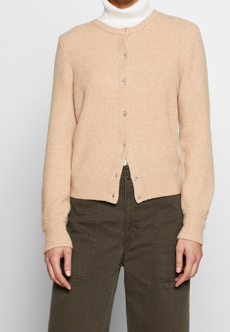Femme portant un cardigan beige boutonné sur un col roulé blanc, associé à un pantalon taille haute vert olive avec des poches plaquées à l'avant.