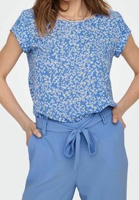 Donna che indossa una maglietta a maniche corte blu floreale infilata nei pantaloni azzurri a vita alta, con una cintura di stoffa annodata, con le mani posate sui fianchi.