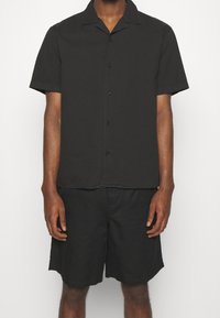 Černá košile s krátkým rukávem a límečkem, se zapínáním na knoflíky, v kombinaci s černými šortkami. Hladký materiál, uvolněný střih.