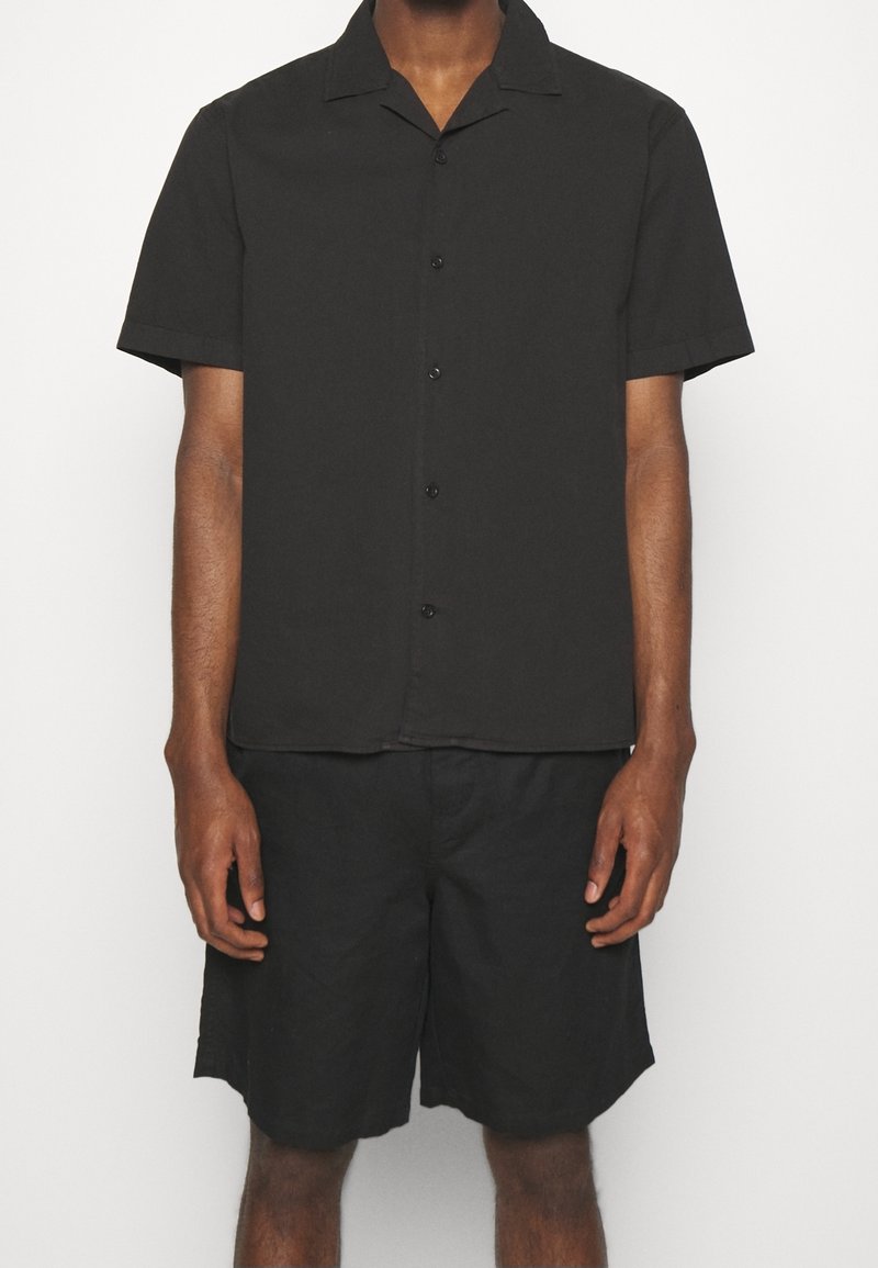 Černá košile s krátkým rukávem a límečkem, se zapínáním na knoflíky, v kombinaci s černými šortkami. Hladký materiál, uvolněný střih.