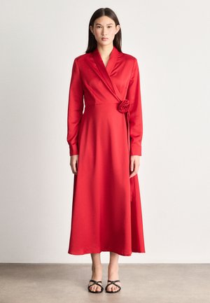 Czerwona sukienka w kształcie litery A z długimi rękawami, dekoltem w serek i różą na wysokości talii. Wykonana z gładkiego materiału, charakteryzuje się zwiewnym krojem.