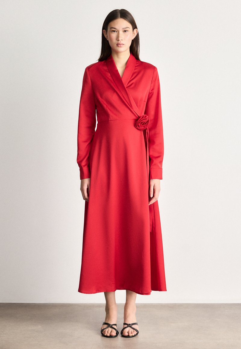 Czerwona sukienka w kształcie litery A z długimi rękawami, dekoltem w serek i różą na wysokości talii. Wykonana z gładkiego materiału, charakteryzuje się zwiewnym krojem.