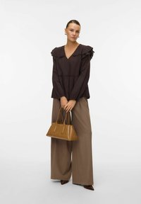 Blouse marron foncé avec des épaules froncées, associée à un pantalon large beige, tenant un petit sac à main marron. Texture lisse et design raffiné.