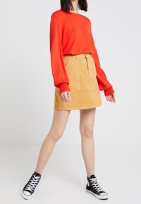 Pull rouge à manches longues en tricot doux, associé à une jupe en denim beige avec des boutons sur le devant et des poches. Baskets montantes noires.