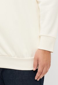 Hvit hettegenser med ribbestrikkede mansjetter og liv, med en jevnt tekstur. Synlig hånd med denim stoff i bakgrunnen.