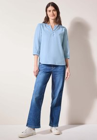 Blusa azzurro chiaro a manica lunga con scollatura a V e chiusura a bottoni. Indossata con jeans blu e sneaker bianche. Texture liscia.