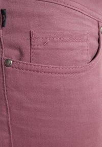 Close-up van een roze spijkerbroekzak met zichtbare stiksels, metalen klinknagel en een klein stoffen label aan de linkerkant.