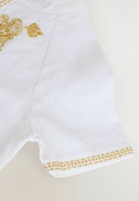 Vêtement en lin blanc avec une broderie dorée intricate et des coutures décoratives le long de l'ourlet. Manches courtes et coupe décontractée.