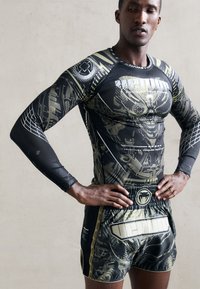 Sort og guld atletisk top med lange ærmer og detaljeret grafisk design, parret med sorte shorts med et fremtrædende logo og accenter.