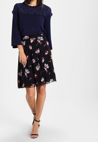 Femme portant un haut en dentelle bleu marine à manches évasées, une jupe fleurie noire longueur genou, et des talons noirs à brides à la cheville, debout devant un fond uni.