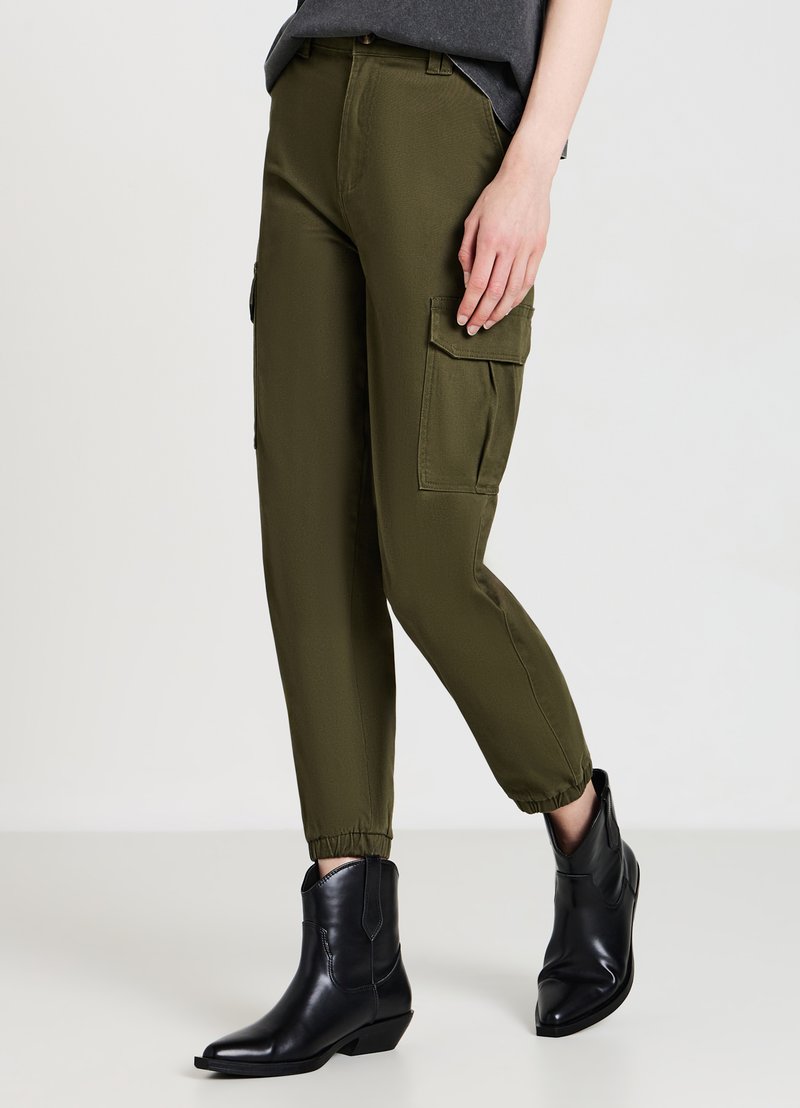 Pantalons cargo vert olive avec des poignets élastiques, dotés de deux poches latérales, associés à des bottines noires en cuir lisse.