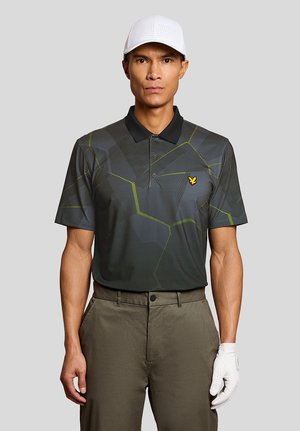 Man met een witte golfpet, donker poloshirt met geometrisch patroon, olijfgroene broek en een witte golfhandschoen aan de rechterhand, staand tegen een grijze achtergrond.
