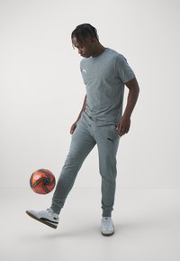 Grå bomullstrøye og joggebukser med svart merkevare, kombinert med hvite joggesko. En oransje og svart fotball er plassert ved føttene.