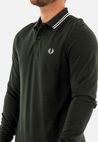 Polo a maniche lunghe verde scuro in tessuto testurizzato, con patta a due bottoni e logo bianco sul petto sinistro. Collo a righe bianche in evidenza.