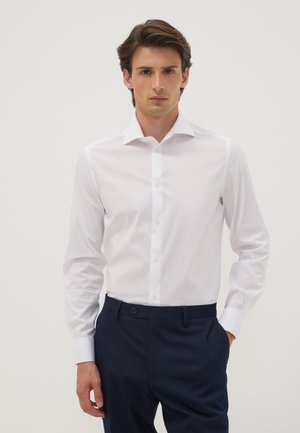 Mann mit braunen Haaren, der ein weißes Hemd und dunkelblaue Hose trägt, steht vor einem einfarbigen hellen Hintergrund.