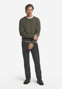 Olivgrön tröja med ribbade ärmslut och nederkant, lager på lager över en vit skjorta. Färgad med mörkgrå jeans och svarta skor. Slim fit-design.