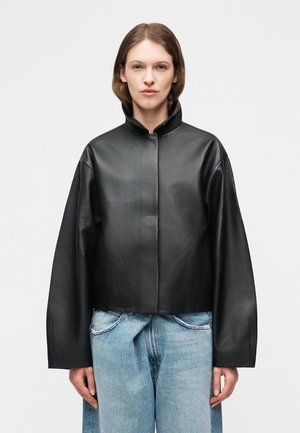 Γυναίκα που φορά υπερμεγέθη μαύρο δερμάτινο μπουφάν με ψηλό γιακά και φαρδιά ανοιχτό μπλε τζιν παντελόνια, στέκεται μπροστά σε απλό φωτεινό φόντο.