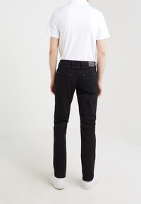 Jeans in denim nero con un design a gamba dritta, dotati di tasche posteriori, una cintura e un'etichetta in pelle sul retro. Indossati con una polo bianca.