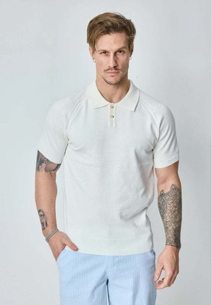 Homme avec des tatouages sur les deux bras portant un polo blanc à manches courtes et un pantalon rayé bleu clair devant un fond uni.