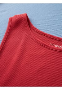 Červený rebrovaný tielkový top s okrúhlym výstrihom položený na svetlomodrom textúrovanom povrchu, zobrazujúci časť značkovej etikety.