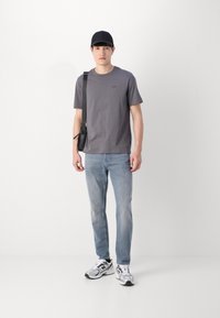 T-shirt en coton gris avec un petit logo, jean bleu clair, casquette noire et baskets grises avec des accents noirs, se tenant devant un fond uni.