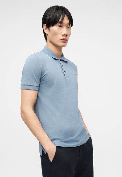 Νεαρός άνδρας με μαύρα μαλλιά, ντυμένος με γαλάζιο κοντομάνικο πόλο και μαύρο παντελόνι, στέκεται με τα χέρια στις τσέπες μπροστά σε λευκό φόντο.