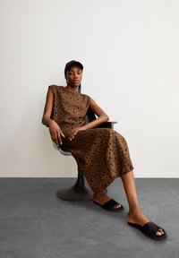 Brown leopard print sleeveless dress, smooth fabric. Black slide sandals. Model seated in a black chair against a neutral backdrop.