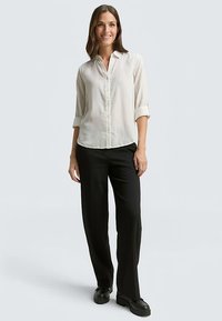 Blouse claire à boutons avec manches retroussées, associée à un pantalon noir ample et des chaussures noires. Tissu lisse, design simple.