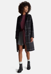 Žena s kudrnatými vlasy, oblečená v tmavém károvaném dlouhém kabátě přes bordó svetr, šedou sukni, bílé ponožky a černé kotníkové boty, stojí a dívá se stranou.