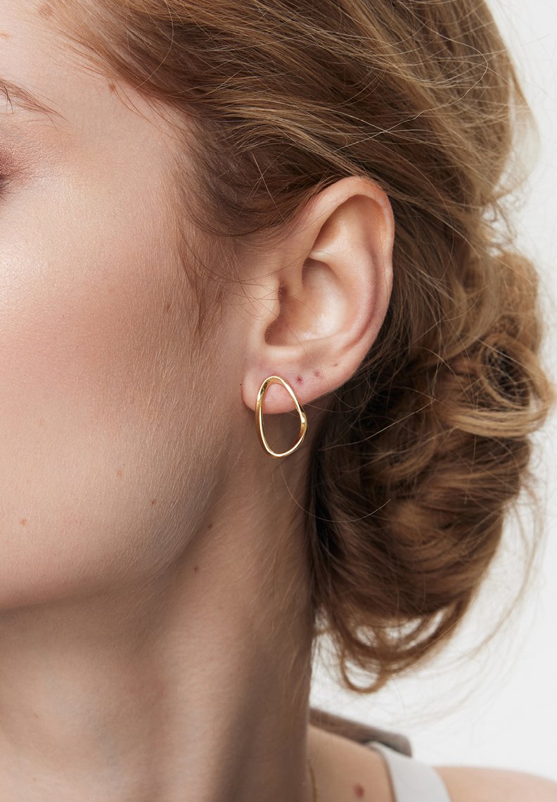 Złota kolczyk w kształcie obręczy o organicznym kształcie. Gładka tekstura, lekko nieregularny kontur i polerowane wykończenie. Subtelny blask na skórze.