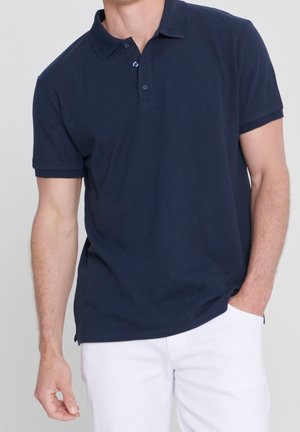 Uomo che indossa una polo blu navy a maniche corte con bottoni e pantaloni bianchi, una mano in tasca, su sfondo semplice.