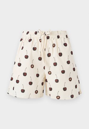 Mujer con una camiseta sin mangas negra, pantalones cortos crema con estampado de cerezas y zapatos planos rojo oscuro, de pie con una mano en el bolsillo.