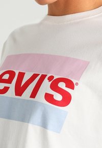 Valkoinen T-paita, jossa on paksu punainen teksti, joka osittain näkyy "Levi's" vaakasuorien vaaleanpunaisten ja vaaleansinisten raitojen päällä.