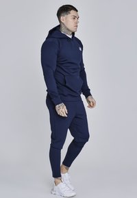 Felpa navy con cappuccio e pantaloni coordinati realizzati in tessuto morbido, con tasca frontale e design aderente. Sneakers bianche con dettagli testurizzati.