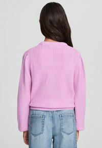 Maglione rosa lavorato a maglia con scollatura rotonda, texture a coste e maniche lunghe, abbinato a jeans di denim blu chiaro con tasche posteriori.