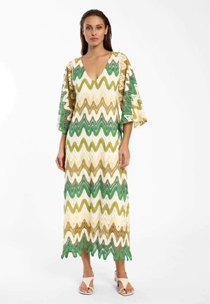 Vrouw die een lange jurk met een golvend patroon draagt in groen, beige en wit met brede mouwen, staand op neutrale sandalen.