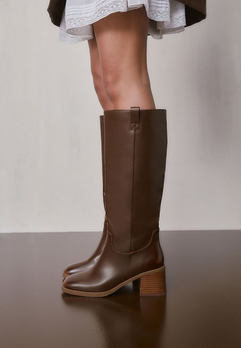 Femme portant des bottes en cuir marron montant jusqu'aux genoux avec des talons blocs, debout sur une surface réfléchissante, visible des genoux jusqu'en bas.