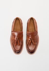 Mocasines de cuero marrón con punta redonda, que presentan borlas en la parte superior, detalles en costura y una suela texturizada.