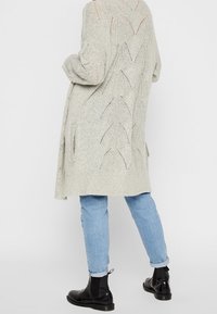 Light gray long cardigan with a textured knit design and openwork detailing. Paired with light blue jeans and black ankle boots.