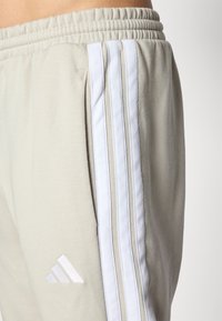 Bežinės sportinės kelnės su baltu elastingu juosmeniu, trys vertikalios baltos juostelės šone ir trikampis logotipas šalia kišenės.