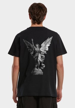 Mann iført svart t-skjorte med detaljert grå englestatue-trykk, med vinger utstrakt og én arm hevet på baksiden.