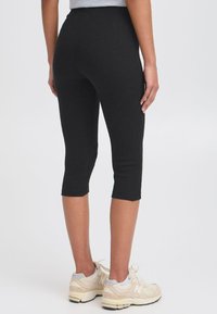Zwarte geribde capri leggings met een slim fit ontwerp, eindigend net onder de knie, gecombineerd met lichtgekleurde sportschoenen.
