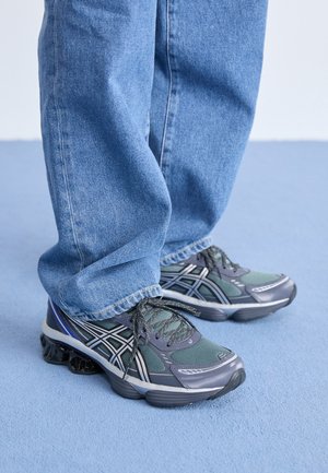 Osoba w niebieskich dżinsach i szaro-zielonych butach do biegania stoi na jasnoniebieskiej podłodze.