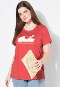 Camiseta roja de manga corta con un gráfico en blanco que dice "en sintonía con la naturaleza". El modelo sostiene un bolso de paja texturizado.