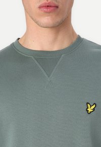 Šviesiai žalias megztinis su grioveliais apykakl�ėje ir kryžminio siuvimo detale, puikuojantis mažuoju juodu ir geltonu paukštelio logotipu ant krūtinės.