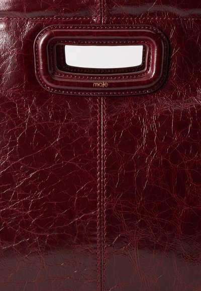 Sac à main en cuir texturé rouge foncé avec poignée découpée rectangulaire et petit logo doré "maje" sous l'ouverture de la poignée.