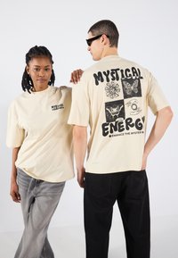 Beige oversized t-skjorter med svarte grafikker, inkludert "MYSTICAL ENERGY" og naturinspirerte design. Modeller bruker avslappede jeans.