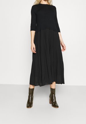 Mujer con una blusa negra de manga larga y una falda midi negra y fluida combinada con botines de tacón marrón oscuro brillantes.