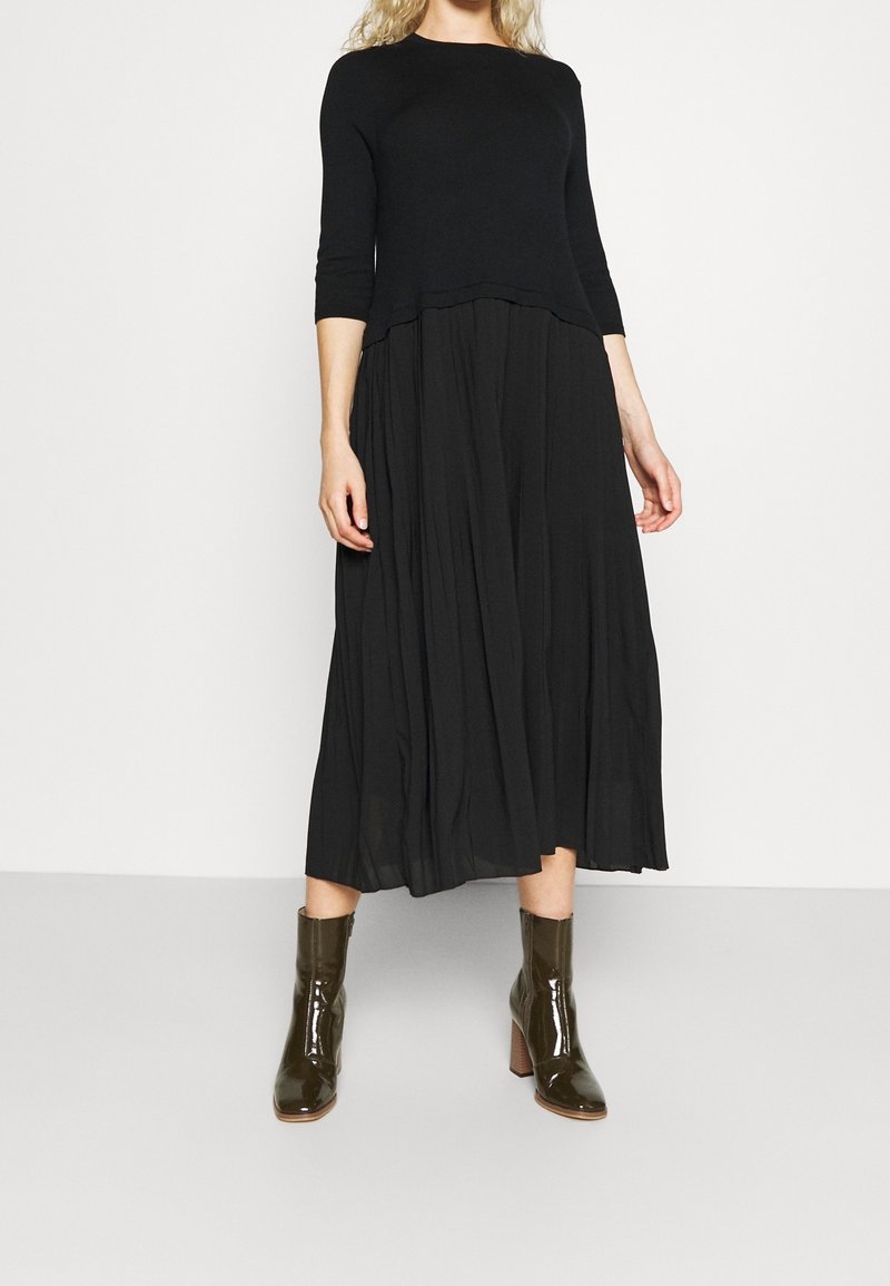 Femme portant un haut noir à manches longues et une jupe midi noire fluide, associée à des bottines à talons brun foncé brillantes.